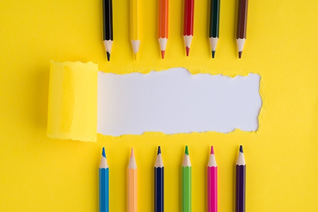 En haut au-dessus de la photo vue de dessus de la rangée de crayons colorés et du papier jaune vif déchiré sur fond blanc avec fond