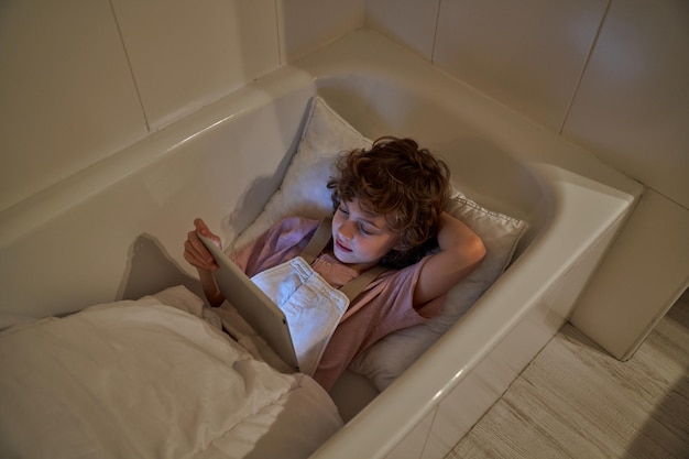 D'en haut, un enfant se repose dans une baignoire avec un oreiller et une couverture tout en regardant une vidéo sur une tablette dans la salle de bain à la maison