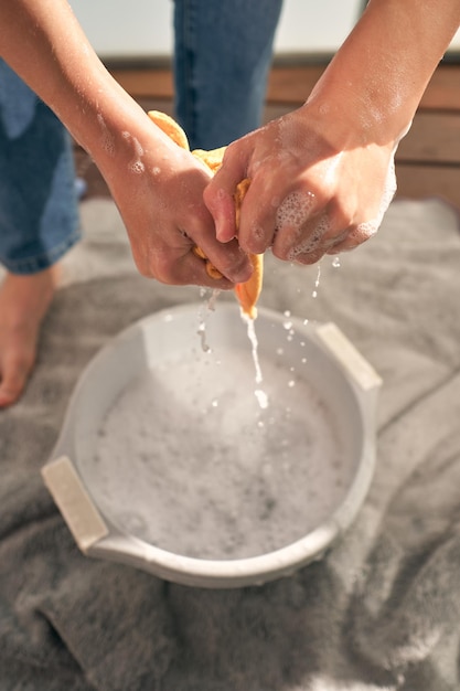 D'en haut d'une personne sans visage en jeans debout sur une serviette et frottant un chiffon sur un bassin blanc avec de l'eau mousseuse tout en faisant la routine domestique à la maison