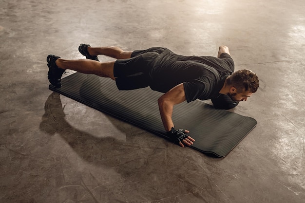 D'en haut, vue arrière d'un athlète masculin en vêtements de sport avec une balle lestée poussant vers le haut en se tenant debout dans une pose de planche dans un gymnase