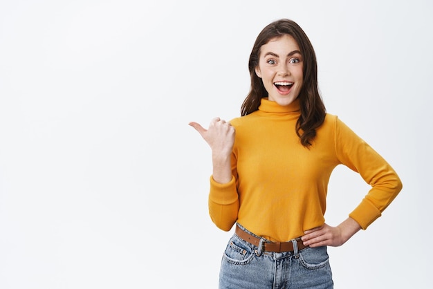 Hé a l'air bien Excité femme souriante pointant le doigt vers la gauche montrant une publicité cool et regardant enthousiaste debout sur fond blanc