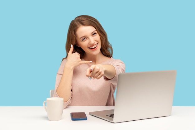 Hé, vous m'appelez Belle charmante femme employée assise sur le lieu de travail avec un ordinateur portable et pointant vers la caméra en gardant la main en forme de téléphone près de la tête prise de vue en studio à l'intérieur isolée sur fond bleu