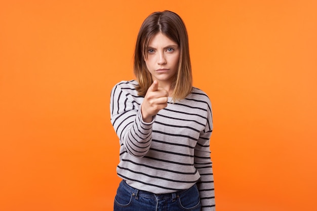 Hé vous Portrait d'une jeune femme mécontente suspecte aux cheveux bruns en chemise rayée à manches longues debout pointant le doigt vers la caméra et blâmant la prise de vue en studio intérieur isolée sur fond orange