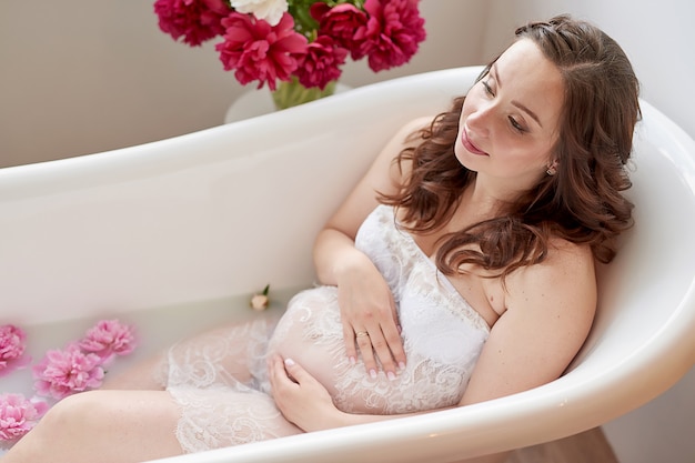 Héhé, femme enceinte dans la cuisine avec des fleurs de pivoines. Maternité, grossesse, concept de bonheur. Fille avec bouquet. Bouquet de pivoines. Fille dans un bain de lait