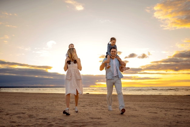 Héhé le long de la plage au coucher du soleil. Jeune belle maman portant sa jolie fille sur les épaules, papa à lunettes porte son bébé. photo de haute qualité