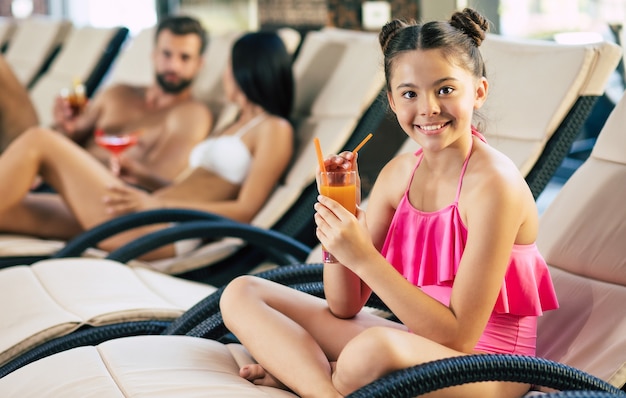 Héhé sur la station. Beau père, belle mère et leur petite fille mignonne sont allongés sur les chaises longues dans le grand centre de spa avec piscine et boissons jus de fruits et cocktails