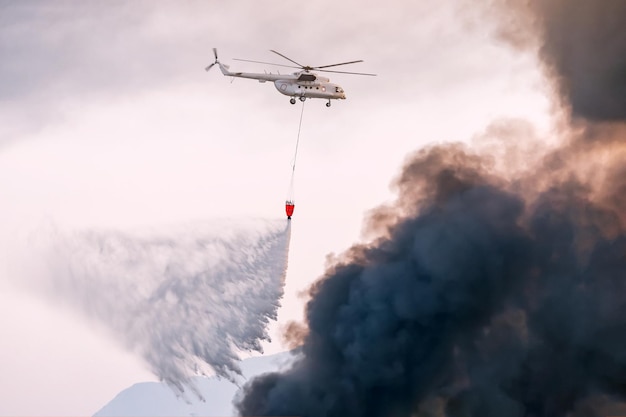 Un hélicoptère d'urgence d'un pompier éteint un incendie et pulvérise de l'eau d'un panier sur une colonne de fumée noire au-dessus d'une ville ou d'un feu de forêt