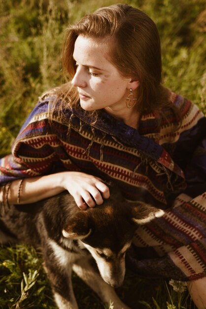 Sur l'herbe de la steppe est assise une fille habillée à la manière des hippies ou boh et caresse le chien