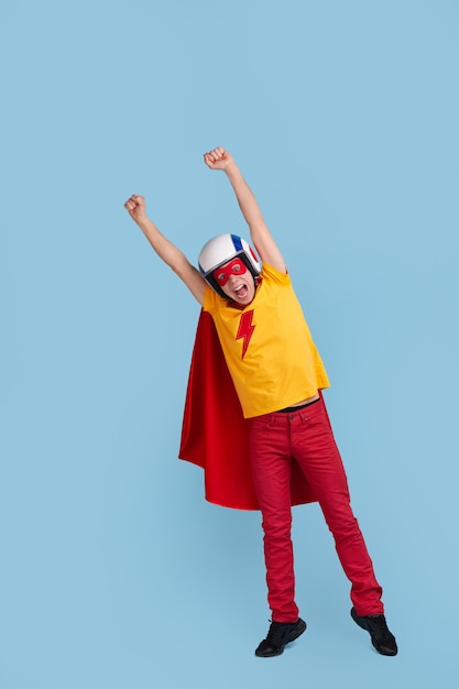 Héros masculin drôle en costume et casque prêt pour le vol debout avec les mains levées sur fond bleu en studio