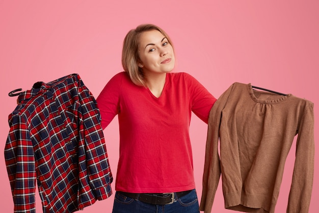 Photo hésitant indécis jeune femme détient deux vêtements, ne peux pas décider quoi porter ce jour, aime faire du shopping et faire des achats, isolé sur mur rose