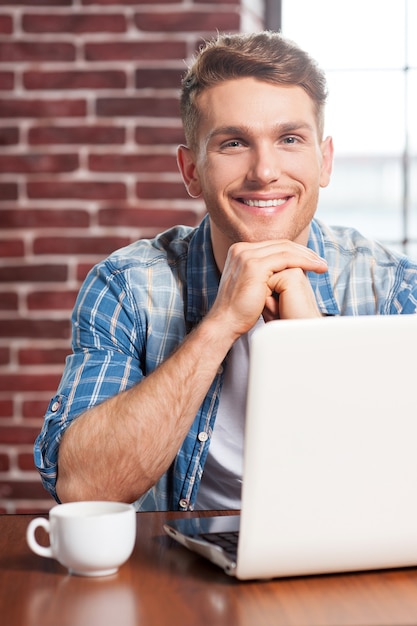 L'heure du café. Beau jeune homme tenant une tasse de café et souriant alors qu'il était assis sur son lieu de travail