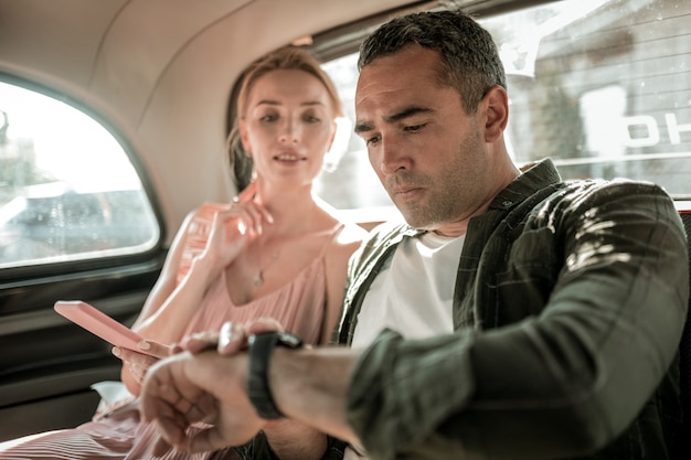 Heure exacte. Homme sérieux réglant sa montre-bracelet vérifiant l'heure sur le smartphone de sa femme assise avec elle dans la voiture.