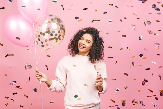 L'heure de la fête! Heureuse belle femme afro-américaine avec un verre de champagne, des ballons et des confettis tombant isolés sur fond rose.