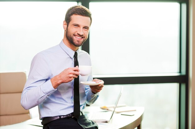 L'heure de la pause-café. Beau jeune homme en chemise et cravate tenant une tasse de café et souriant tout en se penchant au bureau