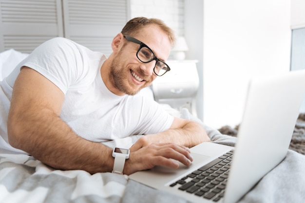 Des heures de travail agréables. Joyeux homme barbu souriant allongé sur le lit et à l'aide de l'ordinateur portable tout en exprimant son intérêt et en travaillant