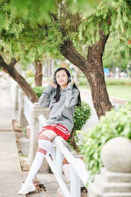 Heureuse adolescente asiatique assis sur la balustrade dans le parc et écouter de la musique dans les écouteurs