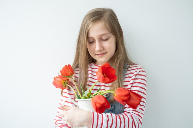 Heureuse adolescente aux cheveux longs tenant un vase avec des tulipes rouges debout contre un mur blanc.