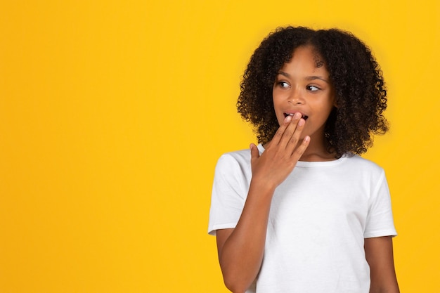 Heureuse adolescente bouclée choquée en t-shirt blanc couvre la bouche ouverte avec la main regarde l'espace libre