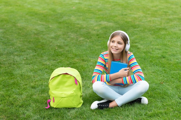 Heureuse adolescente écoutant un cours de formation audio dans un casque assis sur l'herbe après l'école