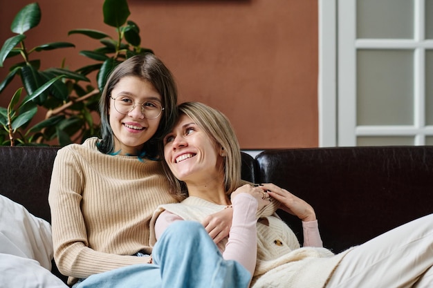 Heureuse adolescente embrassant sa mère et souriant alors qu'ils se reposent sur un canapé dans la chambre