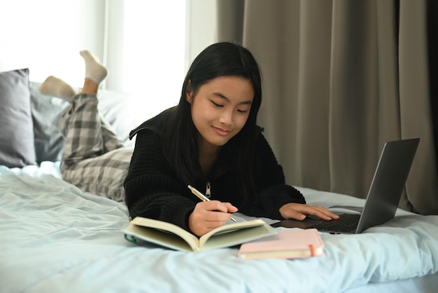 Heureuse adolescente étudiant un cours virtuel d'apprentissage à distance via un ordinateur portable et prenant note sur un ordinateur portable