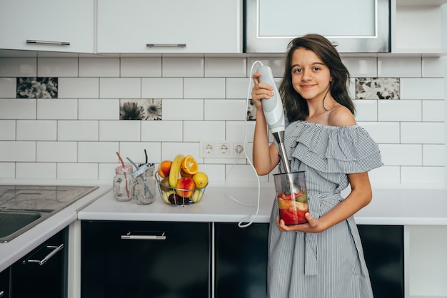 Heureuse adolescente faisant des jus de fruits frais ou un repas dans un mélangeur dans la cuisine.