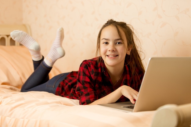 Heureuse adolescente souriante assise sur le lit et utilisant un ordinateur portable