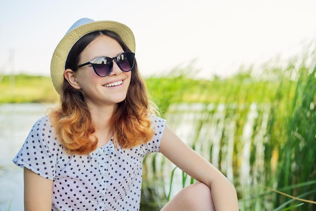 Heureuse adolescente souriante dans des lunettes de soleil chapeau assis sur la rive du lac dans les roseaux, beau fond de nature