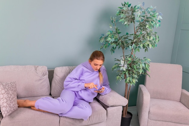 Heureuse adolescente souriante avec son smartphone parcourant le nouveau réseau social communiquant détendu assis sur un canapé à la maison Étude de l'éducation en ligne à distance du concept de maison