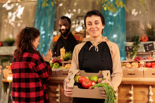 Heureuse agricultrice vendant des produits biologiques sur le marché local, avantages pour la santé des aliments cultivés localement. Vendeur de stand souriant tenant une boîte pleine de fruits et légumes frais de saison.