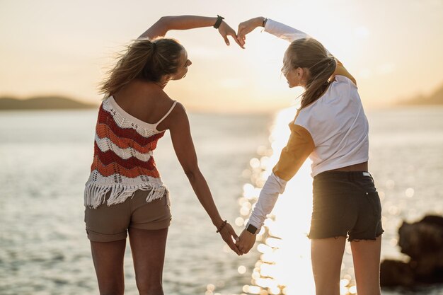 Une heureuse amie passe du temps sur la plage, forme un cœur avec ses mains et profite du coucher du soleil.