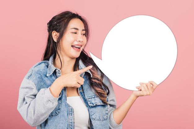 Heureuse asiatique belle jeune femme souriante excitée porter des denims tenir un signe de bulle de parole vide, Portrait femme posant le doigt pointant la bulle pour votre idée de texte, prise de vue en studio isolée sur fond rose