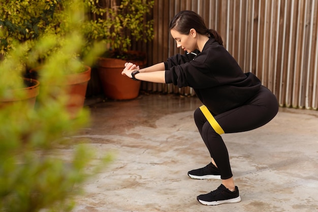 Heureuse athlète européenne du millénaire en tenue de sport accroupie avec un élastique en forme dans la ville en plein air