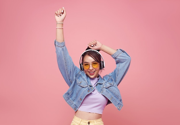 Heureuse belle adolescente asiatique dans des lunettes jaunes, écoutant de la musique dans des écouteurs et dansant sur un mur rose.
