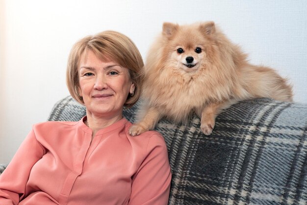 Heureuse belle dame positive, femme âgée âgée assise au canapé dans le salon à la maison avec son animal de compagnie, chien Spitz de Poméranie, petit chiot et souriant. Les gens se soucient, aiment les animaux