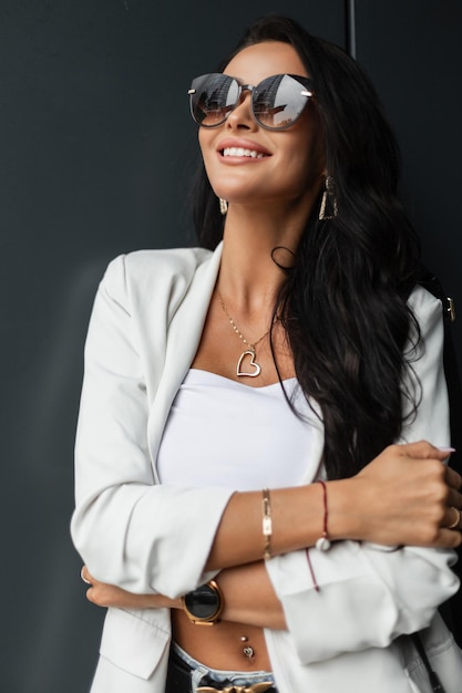 Heureuse belle femme d'affaires souriante avec des lunettes de soleil élégantes dans des vêtements décontractés de mode avec un blazer blanc et une jolie femme urbaine haut avec le sourire profitez