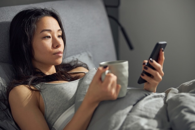 Heureuse belle femme asiatique se détend au lit tout en utilisant un smartphone le matin