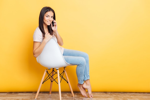 Heureuse belle femme assise sur la chaise et parlant au téléphone sur un mur jaune