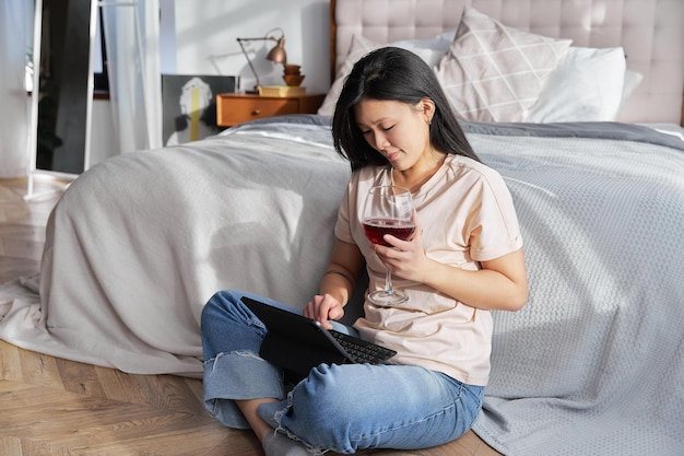Heureuse belle femme dans des vêtements décontractés tenant un verre de vin travaillant sur une tablette numérique assis près d'être