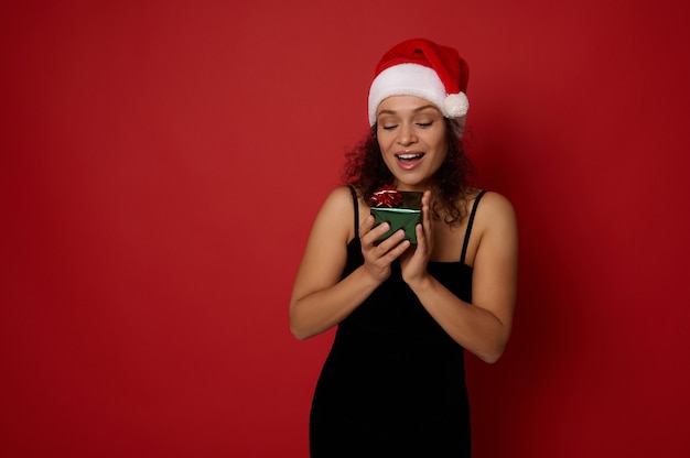 Heureuse belle femme hispanique se sentant heureuse en regardant une petite belle boîte-cadeau de Noël dans ses mains, isolée sur fond rouge avec espace de copie pour l'annonce. Concept de nouvel an et joyeux Noël.