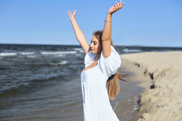 Heureuse, belle femme sur la plage de l'océan debout dans une robe d'été blanche, levant les mains.