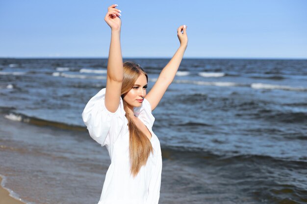 Heureuse, belle femme sur la plage de l'océan debout dans une robe d'été blanche, levant les mains.
