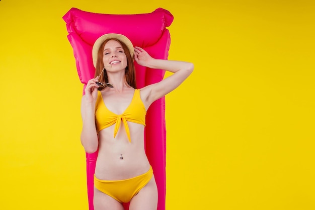 Heureuse belle femme rousse aux cheveux roux en maillot de bain sexy, agitant en soufflant avec un chapeau de paille pour elle-même parce qu'il fait chaud en studio fond jaune. concept de vente d'été.