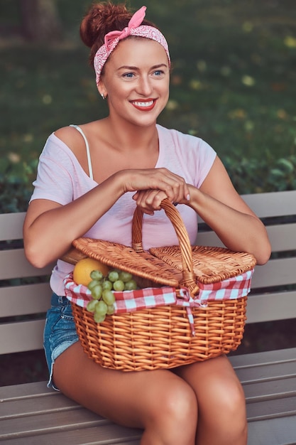 Heureuse belle femme rousse portant des vêtements décontractés tient un panier de pique-nique tout en étant assise sur un banc dans le parc.