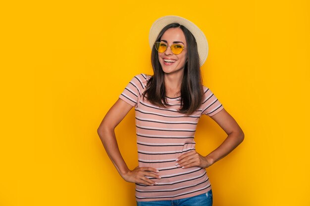 Heureuse belle femme touristique excitée en chapeau et lunettes de soleil pose sur fond jaune et s'amuse