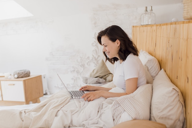 Heureuse belle femme travaillant sur un ordinateur portable assis sur un lit dans une maison moderne et lumineuse