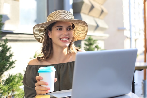 Heureuse belle femme travaillant sur ordinateur portable dans un café de la rue, tenant une tasse en papier.