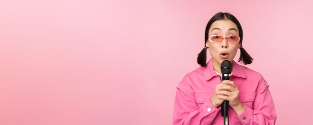 Heureuse belle fille asiatique chantant avec micro à l'aide d'un microphone profitant d'un karaoké posant contre rose st