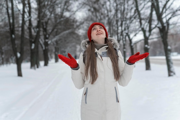 Heureuse belle fille au bonnet rouge en tricot souriant tout en se tenant à bras ouverts et lève les yeux dans le parc d'hiver. Jeune femme sur fond de parc enneigé.