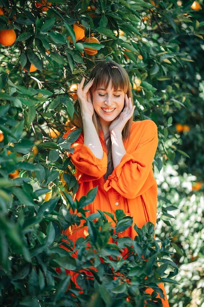Heureuse belle fille en robe orange pose devant la caméra en se tenant la main près du visage dans le jardin orange